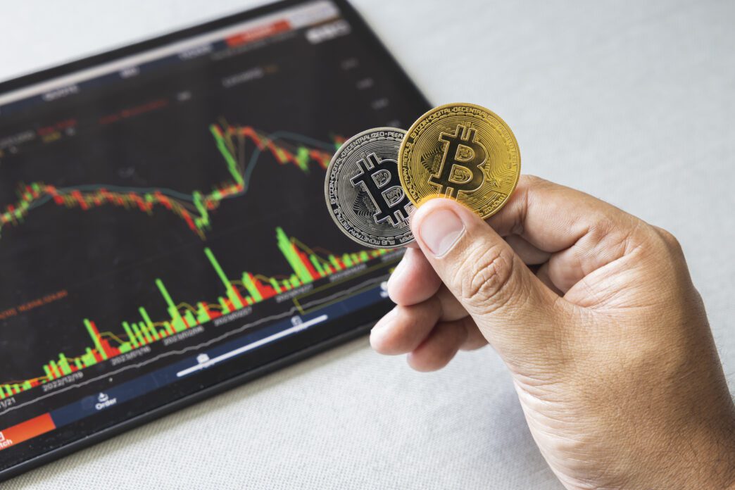 Hand holding one gold and one silver Bitcoin coin over a tablet displaying a red and green trading chart.