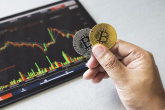 Hand holding one gold and one silver Bitcoin coin over a tablet displaying a red and green trading chart.