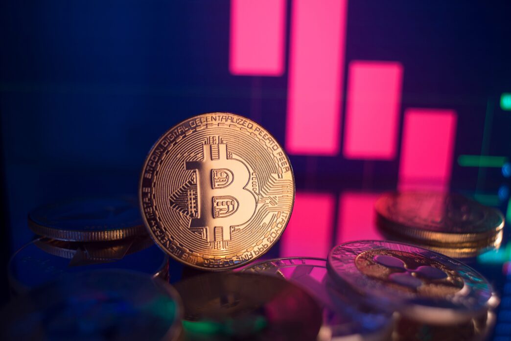 A golden physical Bitcoin coin stands on a dark surface in front of a digital screen showing a red, sharply declining financial candlestick chart.