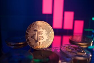 A golden physical Bitcoin coin stands on a dark surface in front of a digital screen showing a red, sharply declining financial candlestick chart.
