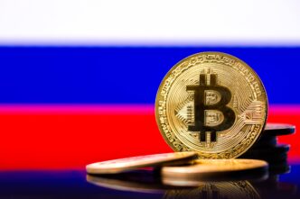A golden physical Bitcoin coin prominently displayed against the blurred white, blue, and red background of the Russian flag.
