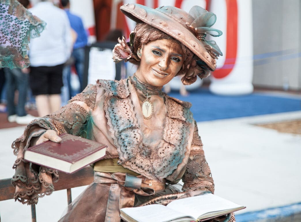 Female street performer, painted bronze and turquoise, smiling while holding out a book in Miami.