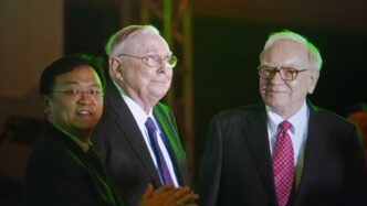 Warren Buffett and Charlie Munger stand with a third man at the Berkshire Hathaway meeting.