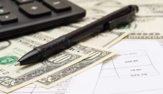Black pen resting on US dollar bills and paper financial documents, with a calculator in the background.