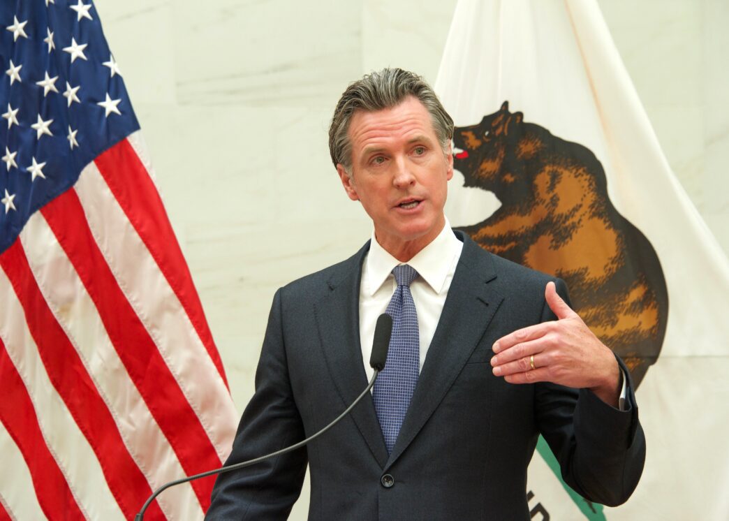California Governor Gavin Newsom speaks at a podium, standing between the American flag and the California state flag.