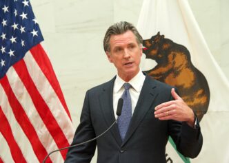 California Governor Gavin Newsom speaks at a podium, standing between the American flag and the California state flag.
