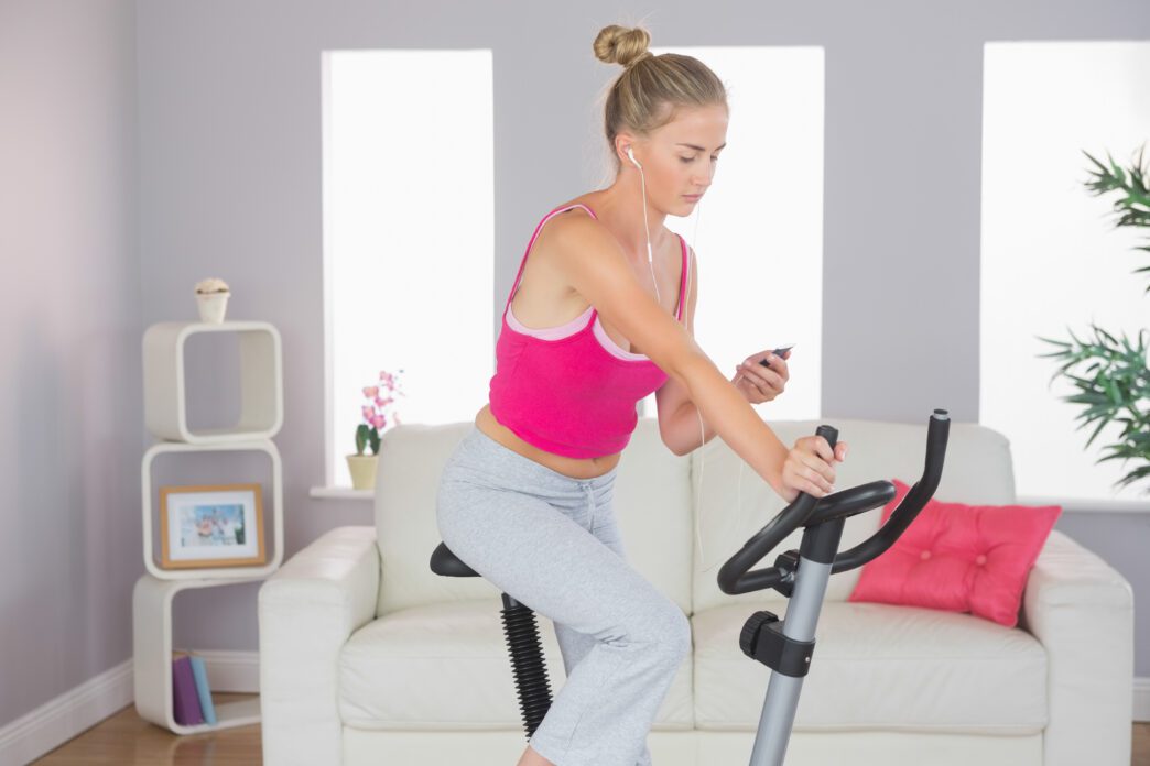 Blonde woman exercising on a stationary bike indoors, listening to music on her smartphone.
