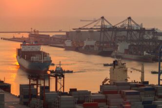 Aerial view of a busy commercial harbor with large cargo ships and cranes silhouetted against a sunrise.