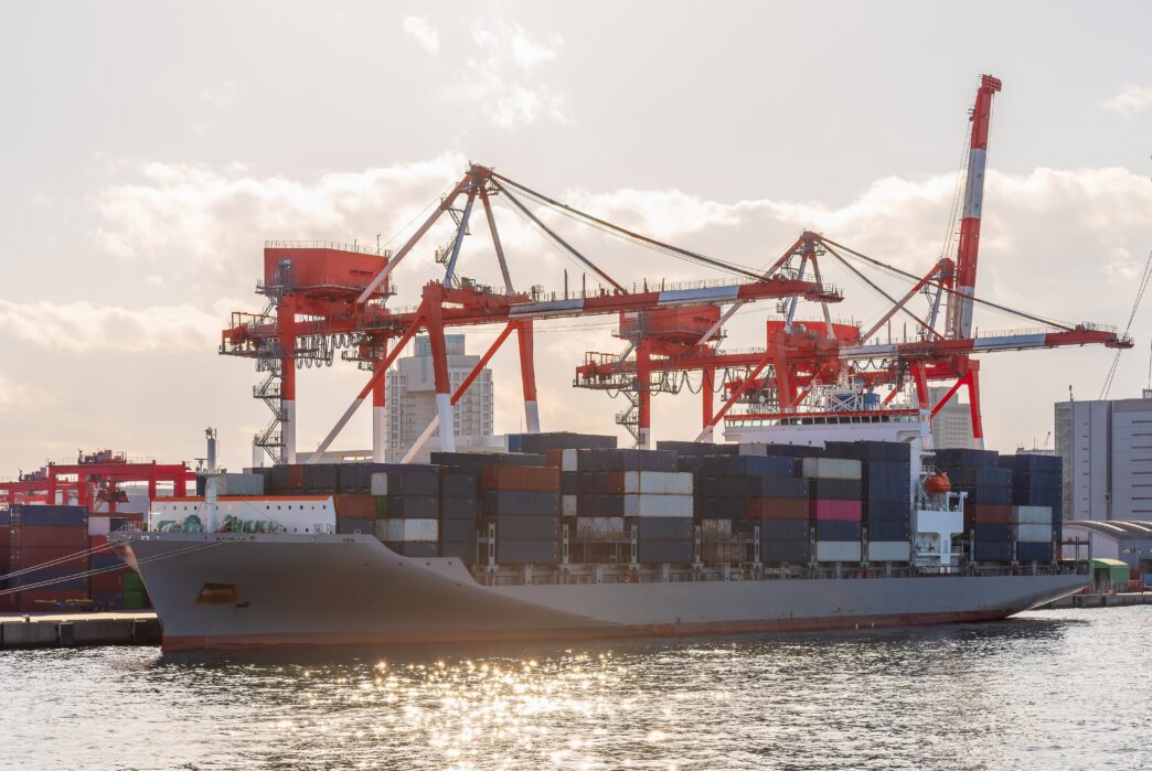 Large container ship docked at a port in Osaka, Japan, being loaded by massive red and white gantry cranes under a bright sky.