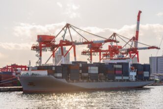 Large container ship docked at a port in Osaka, Japan, being loaded by massive red and white gantry cranes under a bright sky.