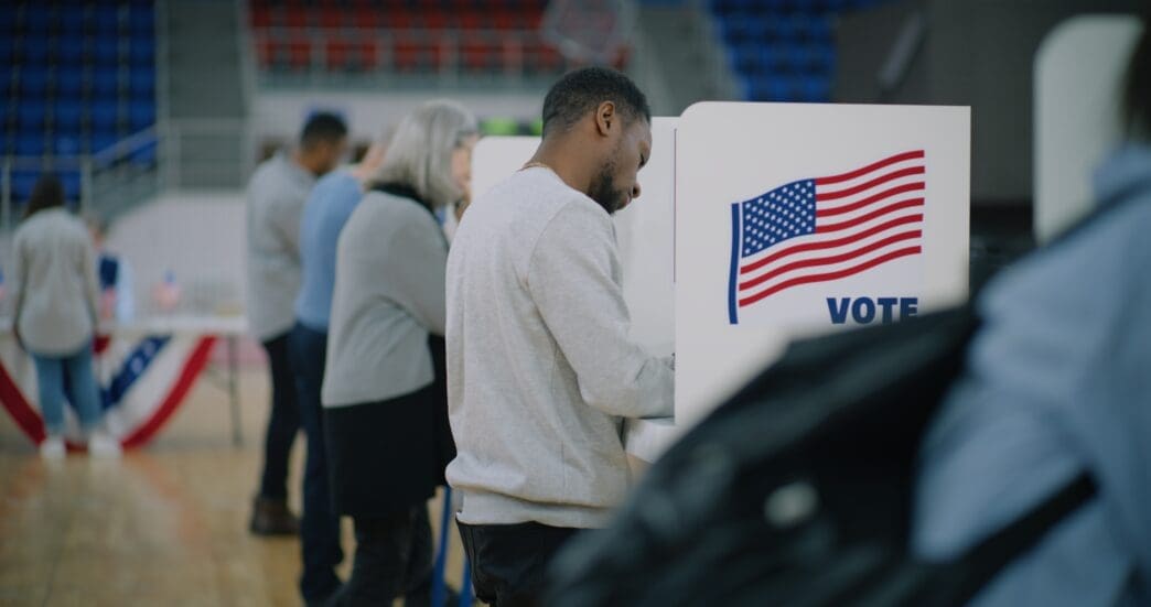 A diverse group of American citizens, including a man in the foreground, cast their votes at booths on Election Day.