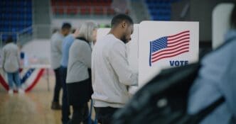 A diverse group of American citizens, including a man in the foreground, cast their votes at booths on Election Day.