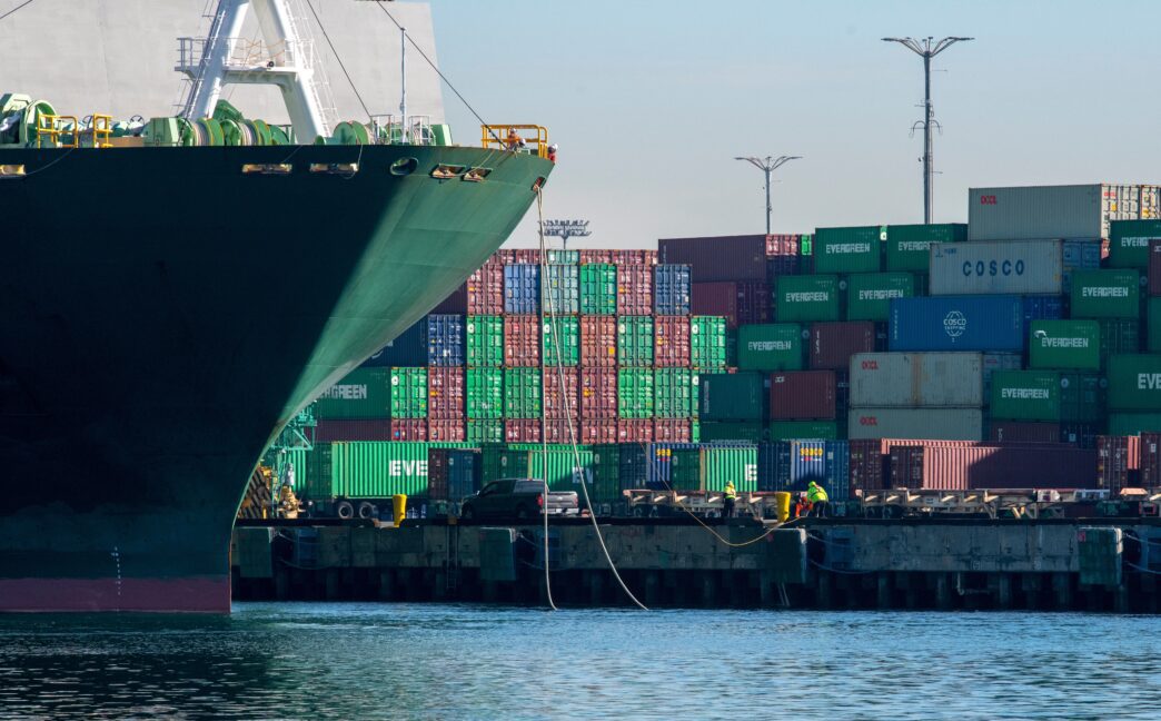Large container ship docked at the Port of Los Angeles, with dockworkers securing ropes and a massive stack of colorful shipping containers in the background.