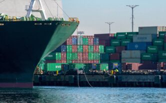 Large container ship docked at the Port of Los Angeles, with dockworkers securing ropes and a massive stack of colorful shipping containers in the background.