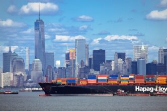 A large Hapag-Lloyd container ship sailing past the Manhattan skyline, including One World Trade Center.