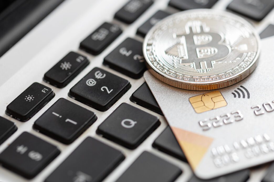 Silver Bitcoin coin resting on a credit card and laptop keyboard, linking crypto to digital payment.