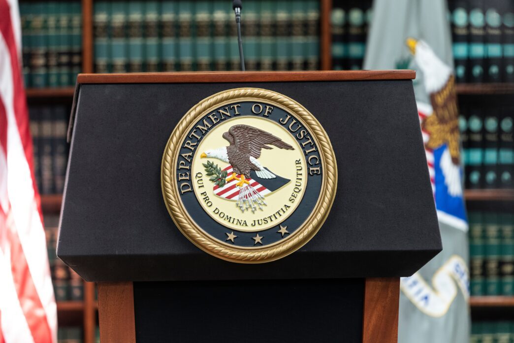 The gold seal of the U.S. Department of Justice centered on a black podium, with flags and books in the background.