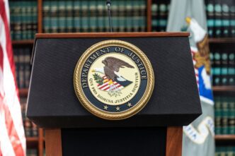 The gold seal of the U.S. Department of Justice centered on a black podium, with flags and books in the background.