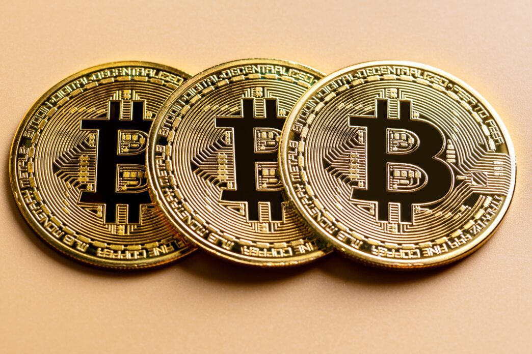 Three shiny gold Bitcoin cryptocurrency coins lined up on a smooth beige surface.