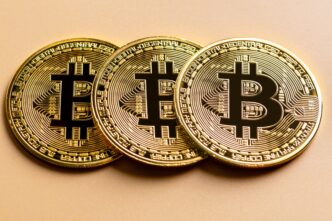 Three shiny gold Bitcoin cryptocurrency coins lined up on a smooth beige surface.