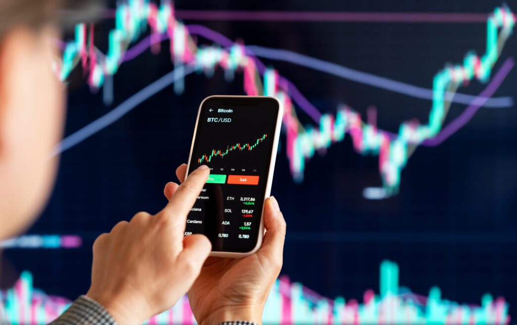 A female investor holds a smartphone displaying a cryptocurrency trading app with Bitcoin (BTC) charts and a green 'Buy' button.