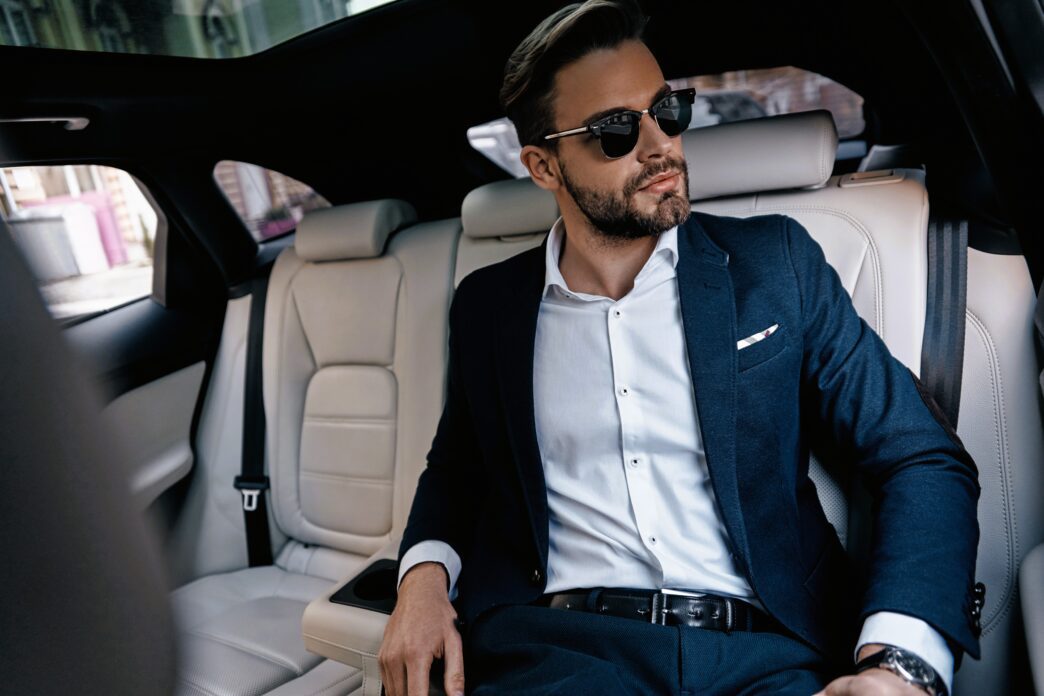 Stylish man in a dark suit and sunglasses sitting in the back seat of a luxury car.