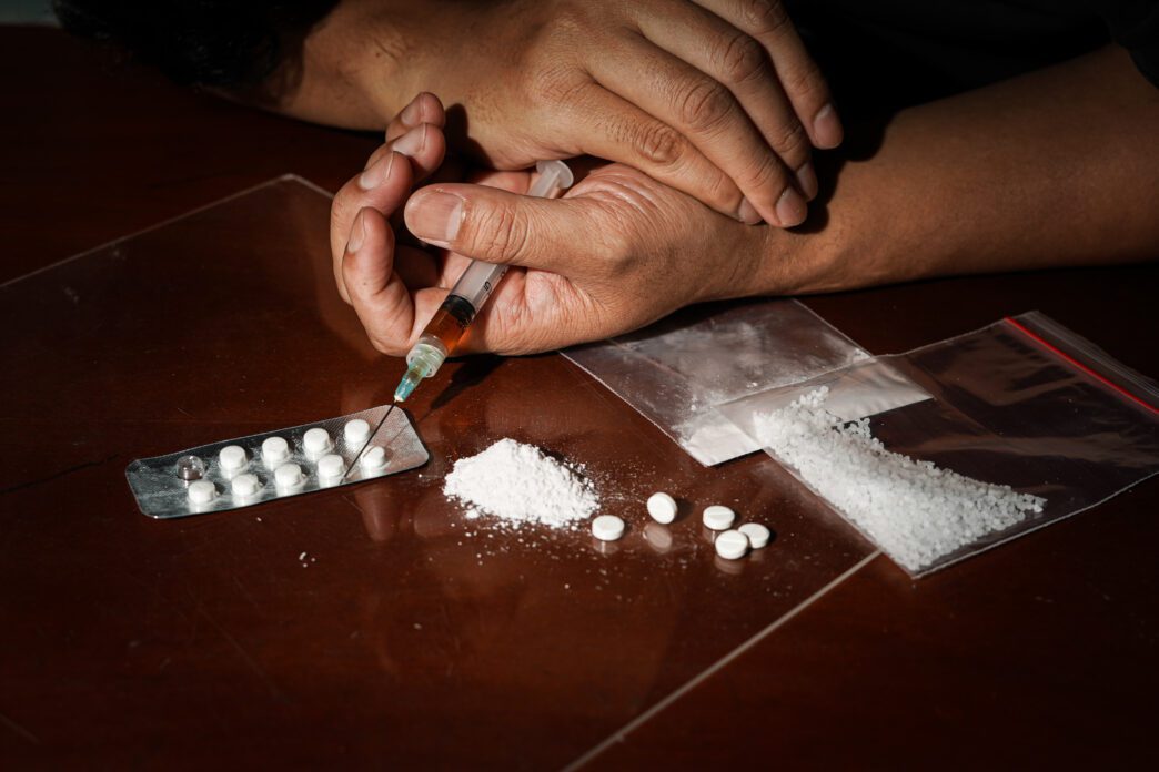 Hands holding a syringe near pills, powder, and plastic bags on a dark table, symbolizing drug abuse.
