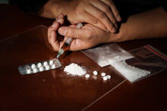 Hands holding a syringe near pills, powder, and plastic bags on a dark table, symbolizing drug abuse.