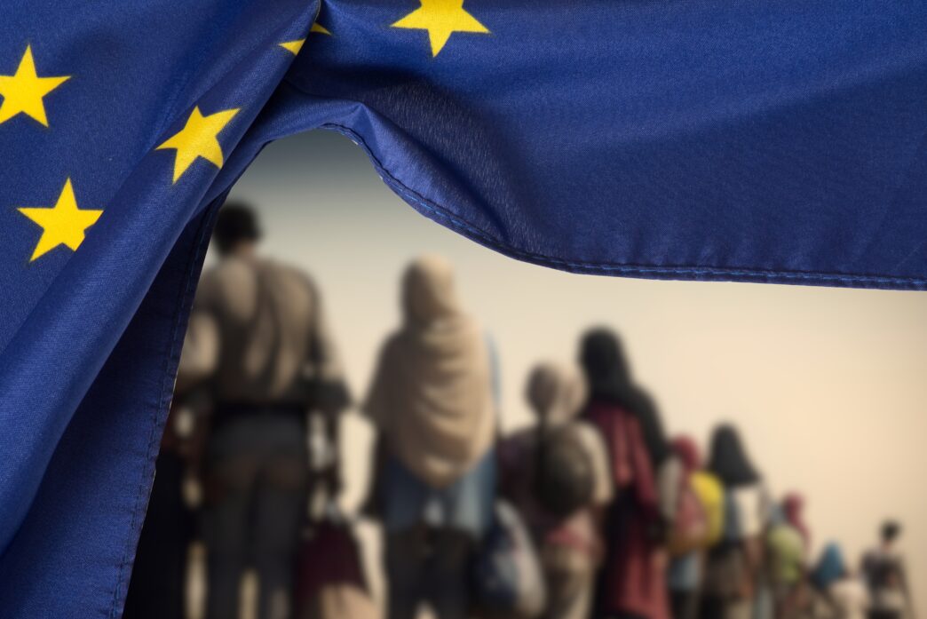 EU flag partially framing a blurred group of migrants walking towards the background.