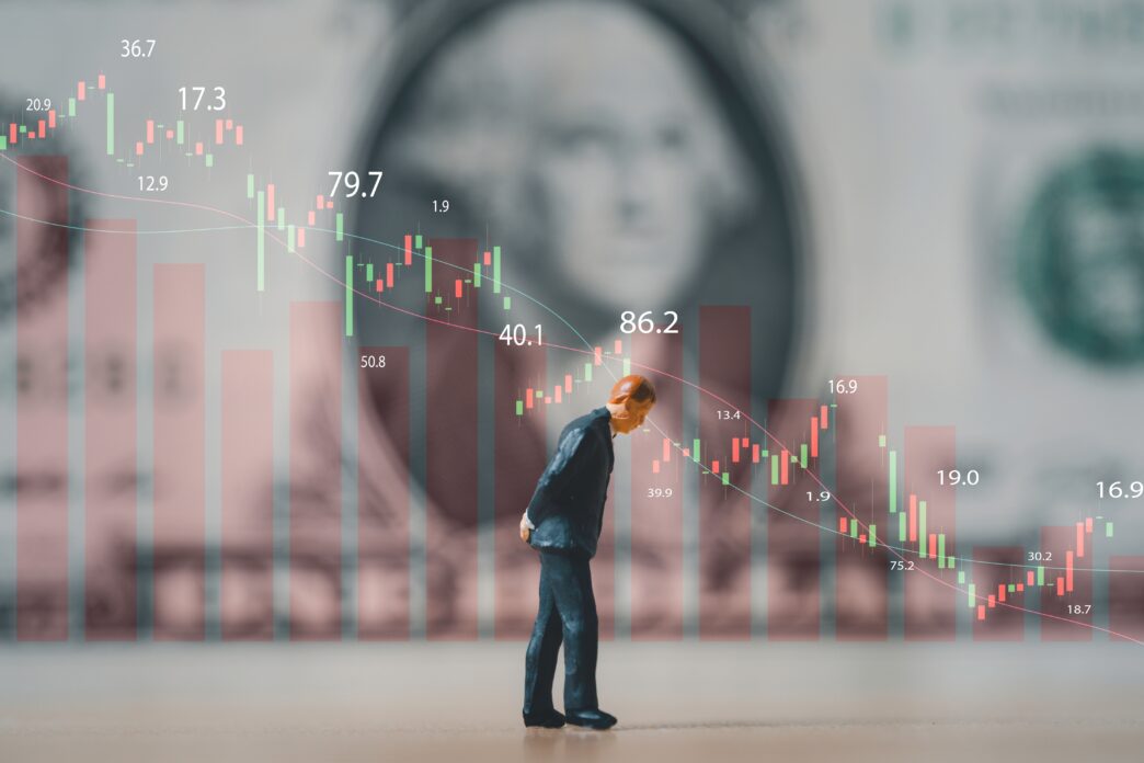 Miniature businessman looking down in front of a falling stock market graph and a blurred dollar bill.