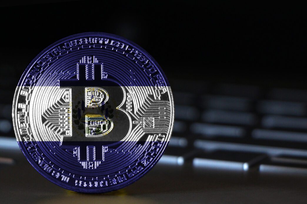 A physical Bitcoin coin, displaying the flag of El Salvador, rests on a dark laptop keyboard.