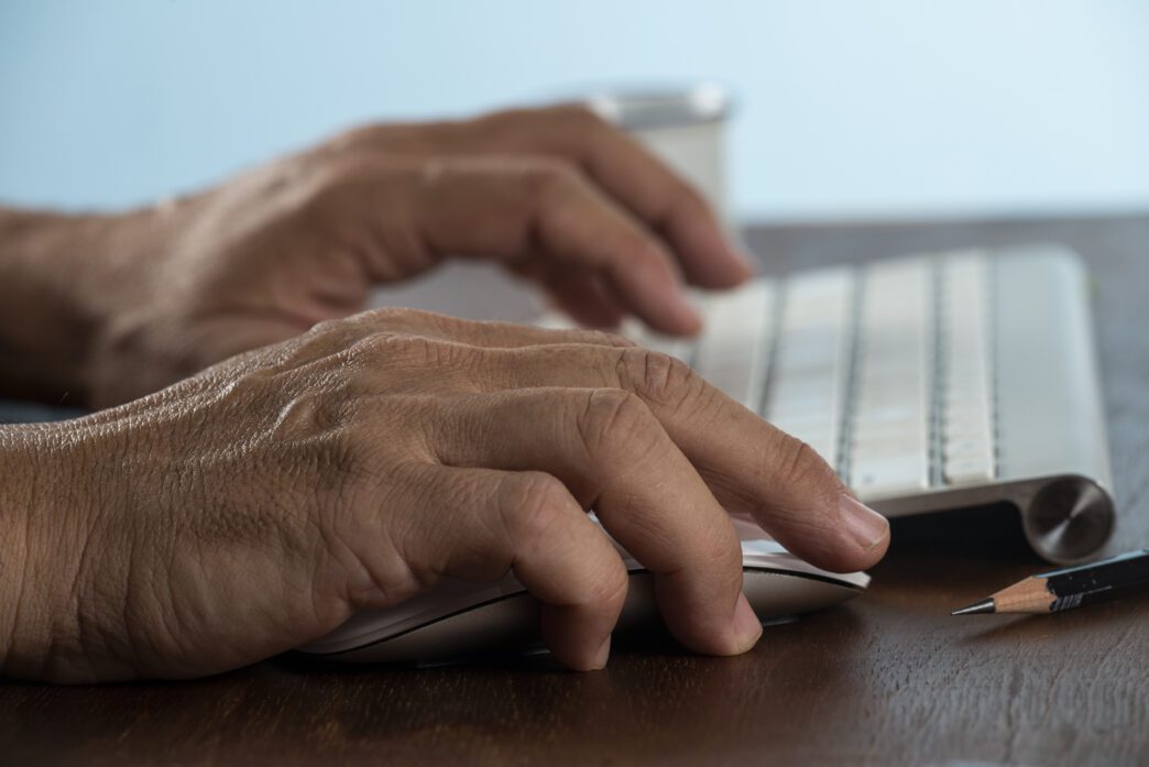Close-up of aging hands using a modern computer keyboard and mouse on a wooden desk.