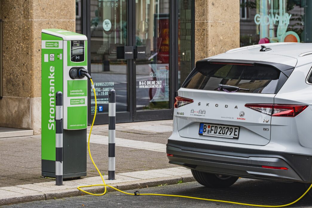 A white Skoda Enyaq iV is charging at a green and white "Stromtanke" electric vehicle charging station on a city street.