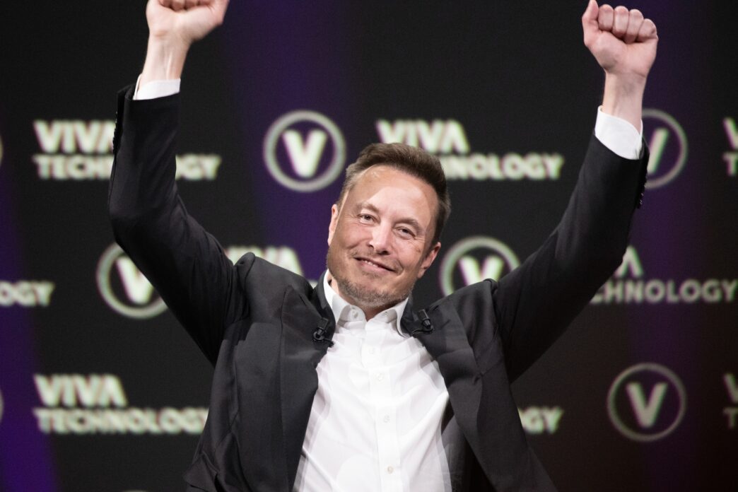 Elon Musk, wearing a suit, raises both arms in a triumphant gesture at a technology conference.