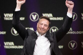 Elon Musk, wearing a suit, raises both arms in a triumphant gesture at a technology conference.
