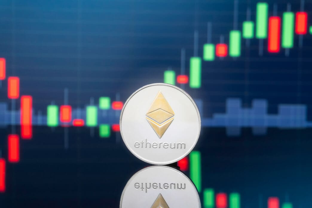 A physical silver Ethereum coin stands on a reflective surface against a financial chart with red and green candlesticks.