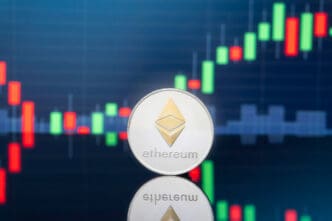 A physical silver Ethereum coin stands on a reflective surface against a financial chart with red and green candlesticks.