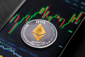 Physical Ethereum coin resting on a smartphone screen displaying a red and green trading chart.