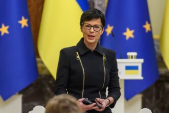 Slovenian diplomat Marta Kos, wearing a black blazer with gold zippers and glasses, speaks at a press event.