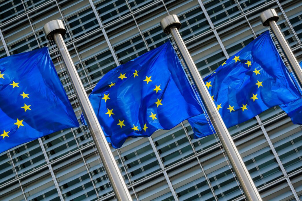 Three blue European Union flags with yellow stars fly in front of the modern, glass-and-steel Berlaymont building.