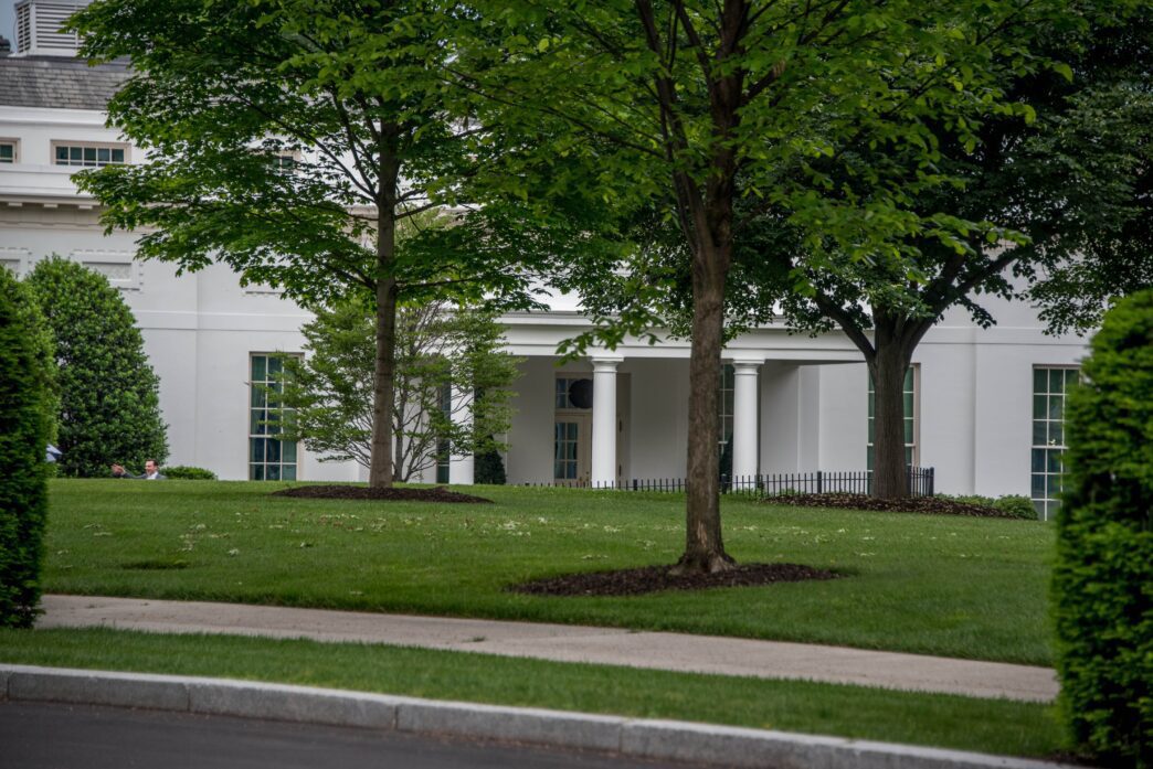 Exterior view of the White House West Wing portico and facade, partially obscured by green trees and lawn.