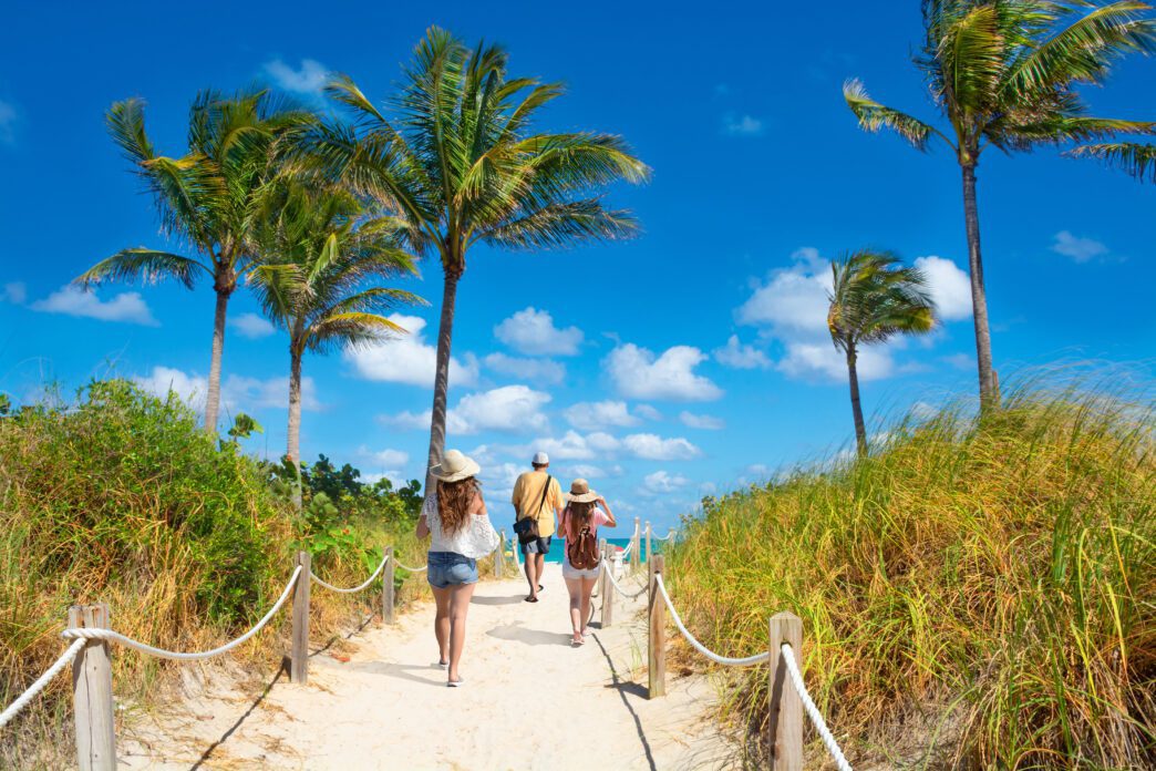 Three people walk away from the camera on a sandy beach path lined with ropes, palm trees, and tall grass, heading toward the ocean under a blue sky.