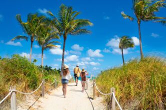 Three people walk away from the camera on a sandy beach path lined with ropes, palm trees, and tall grass, heading toward the ocean under a blue sky.