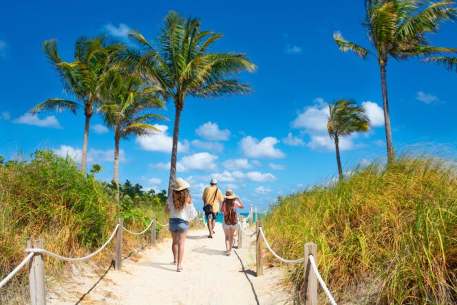 Three people walk away from the camera on a sandy beach path lined with ropes, palm trees, and tall grass, heading toward the ocean under a blue sky.