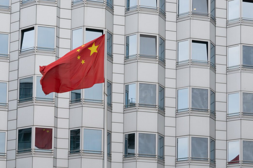 The red flag of China, featuring five yellow stars, flies from a modern, multi-story building with repeating square windows.