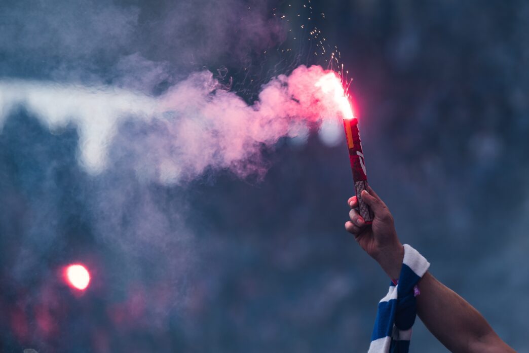 A fan's arm holding a brightly burning red smoke flare over a crowded stadium.