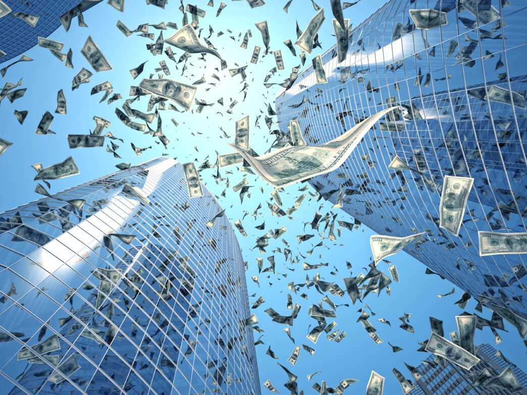 US dollar bills raining down between two tall, glass skyscrapers under a blue sky.