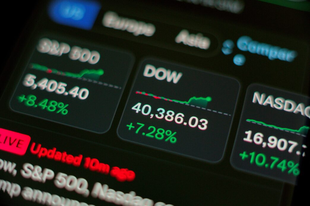 Close-up of a digital screen showing stock market indices (S&P 500, DOW, NASDAQ) with high percentage gains.