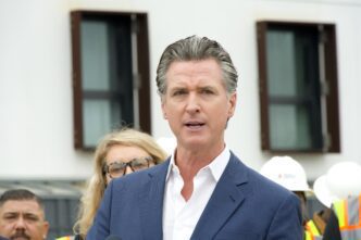 Close-up of Governor Gavin Newsom speaking at an outdoor press event in front of a modern building.