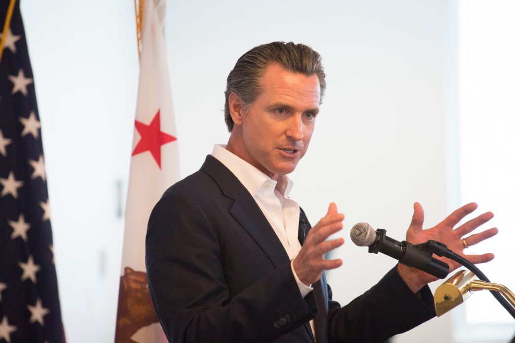 Governor Gavin Newsom speaking at a podium, gesturing with his hands, flanked by US and California flags.